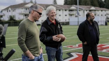 Coach Mac holding a football standing on football field with past students.