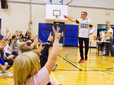 Students participate in a Science World presentation in a school gymnasium by raising their hands to answer questions.