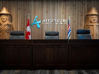 Abbotsford School District boardroom with a dark wood council-style desk and three black chairs in the foreground. A wood slat feature wall displays the Abbotsford School District logo and name, flanked by the Canadian and British Columbia flags. Two large carved wooden totem-style figures stand on either side of the room, creating a formal meeting setting.